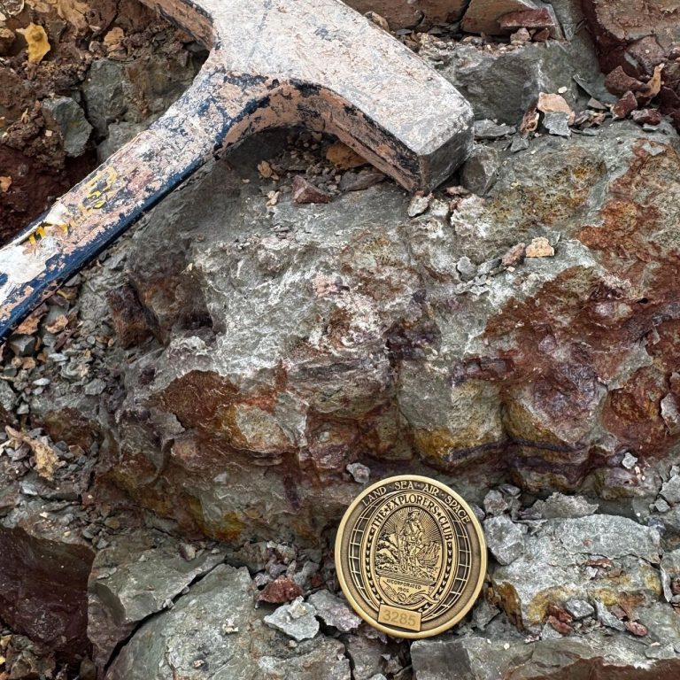 Rock hammer and an Explorers Club challenge coin for scale, resting on grey and red rock at an Algarve outcrop.