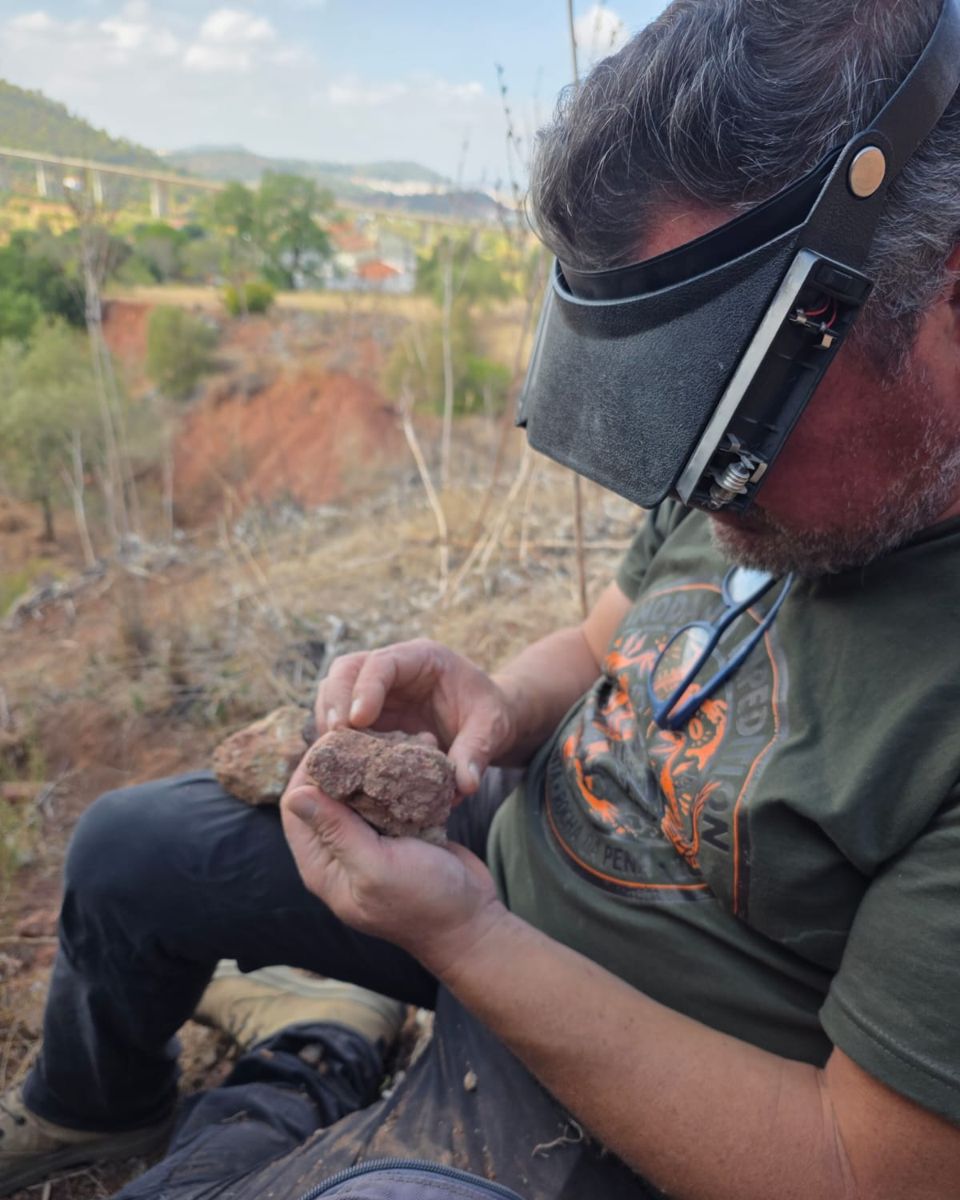 Bruno Camilo, wearing a magnifying visor, turns over a piece of reddish rock while sitting on a hillside