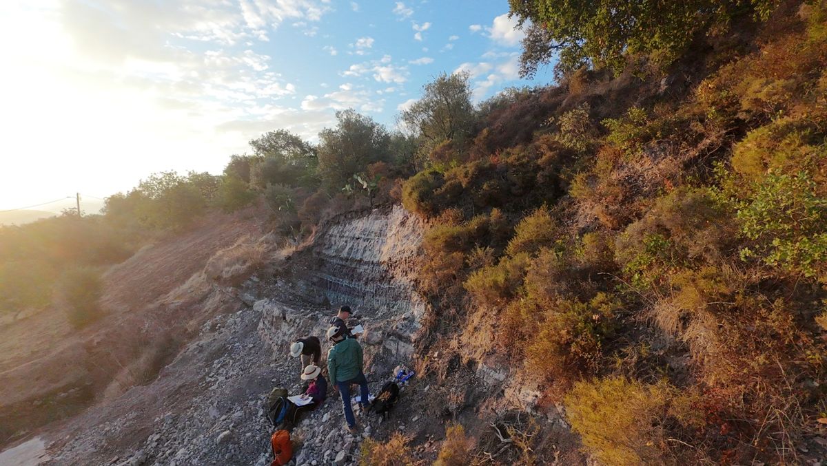 Members of the DinoDawn team with notebooks at the foot of a cut of grey and white banded rock in the Algarve hills