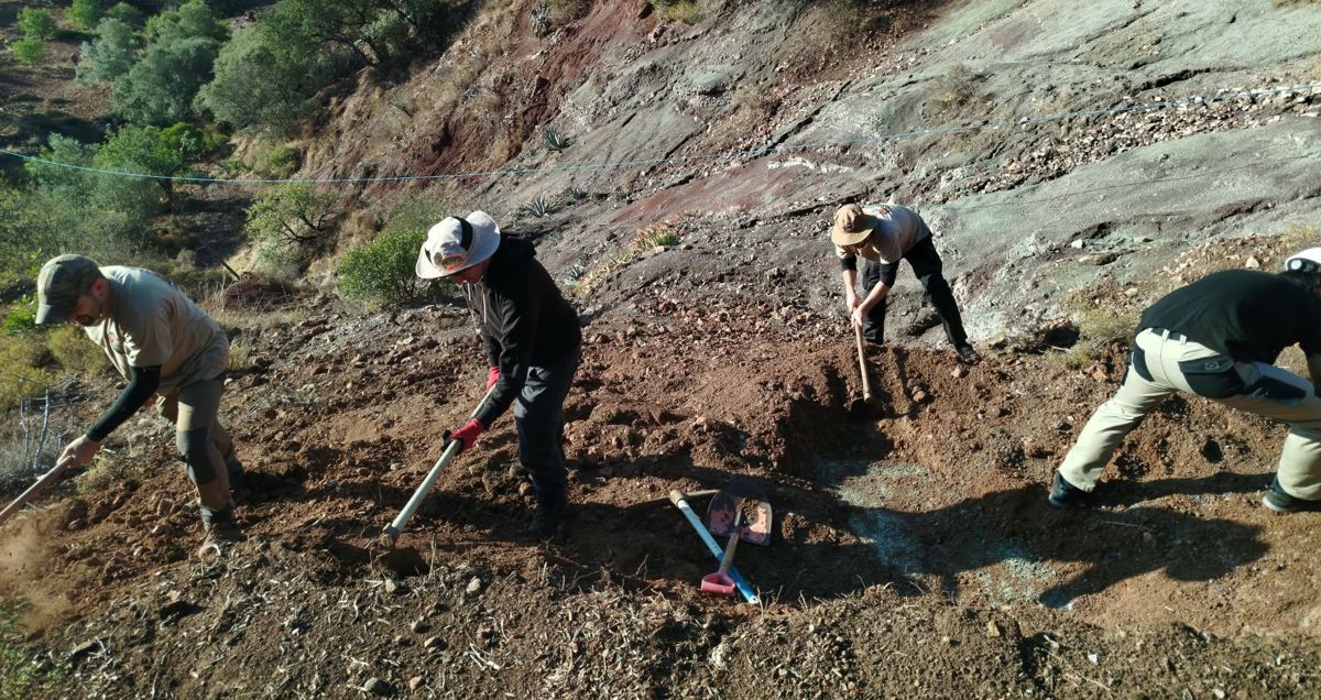 4 team members with hoes clearing reddish soil from a slope above a pale gray rock face