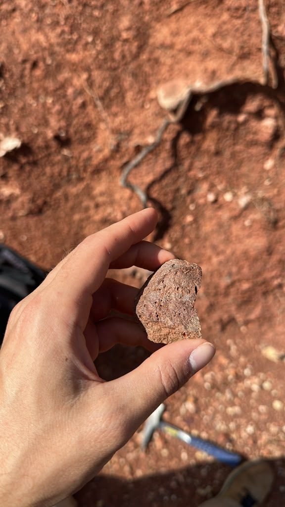 A hand holding a pitted reddish rock sample above red soil, with a rock hammer on the ground below