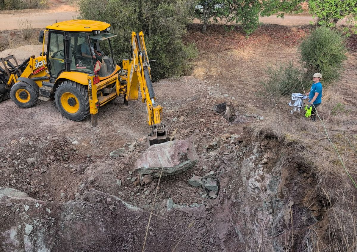 A yellow backhoe excavator lifting a large pink and gray block of rock, with a team member watching from the edge