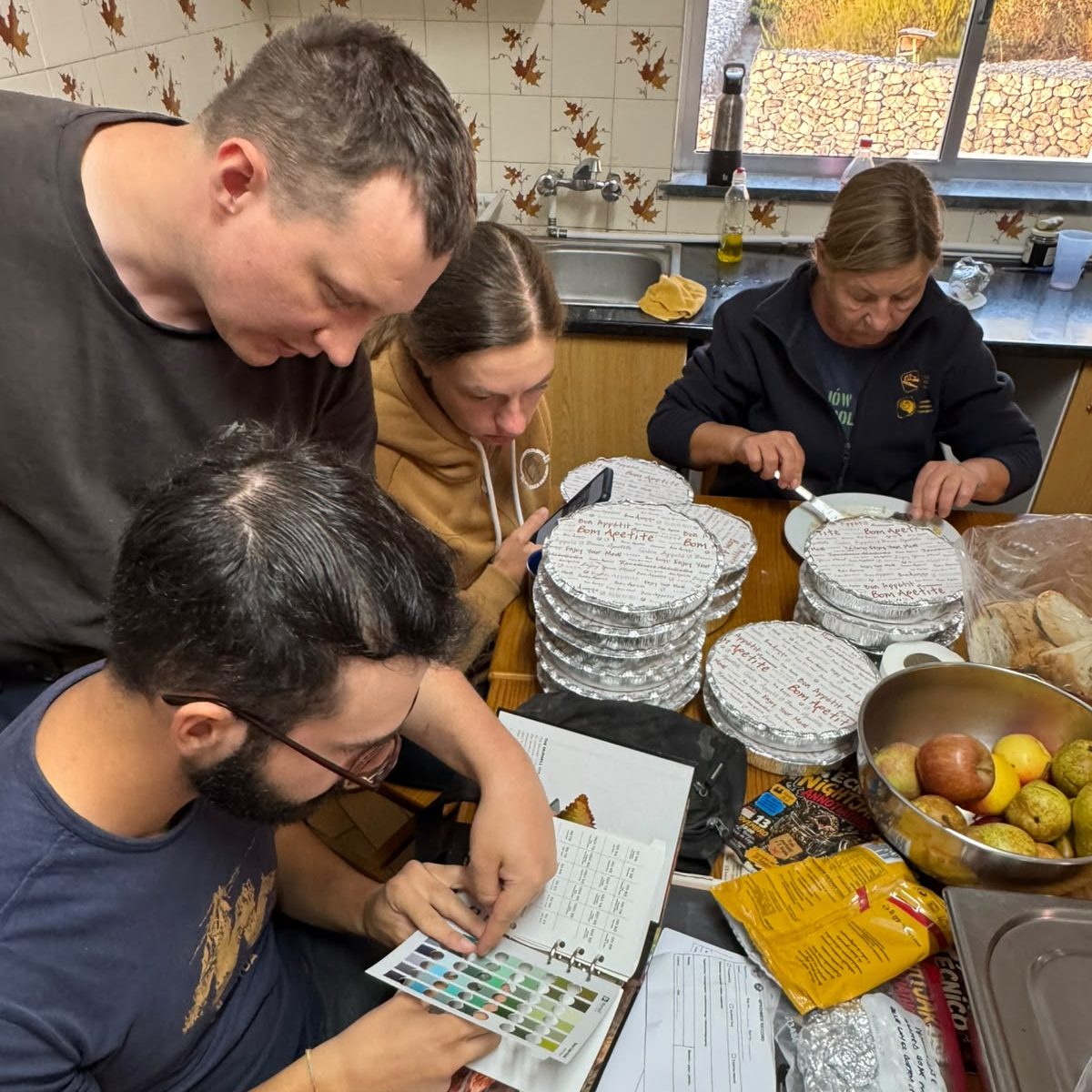 Team members at a kitchen table, one holding a color chart against a binder, with dinner trays stacked nearby