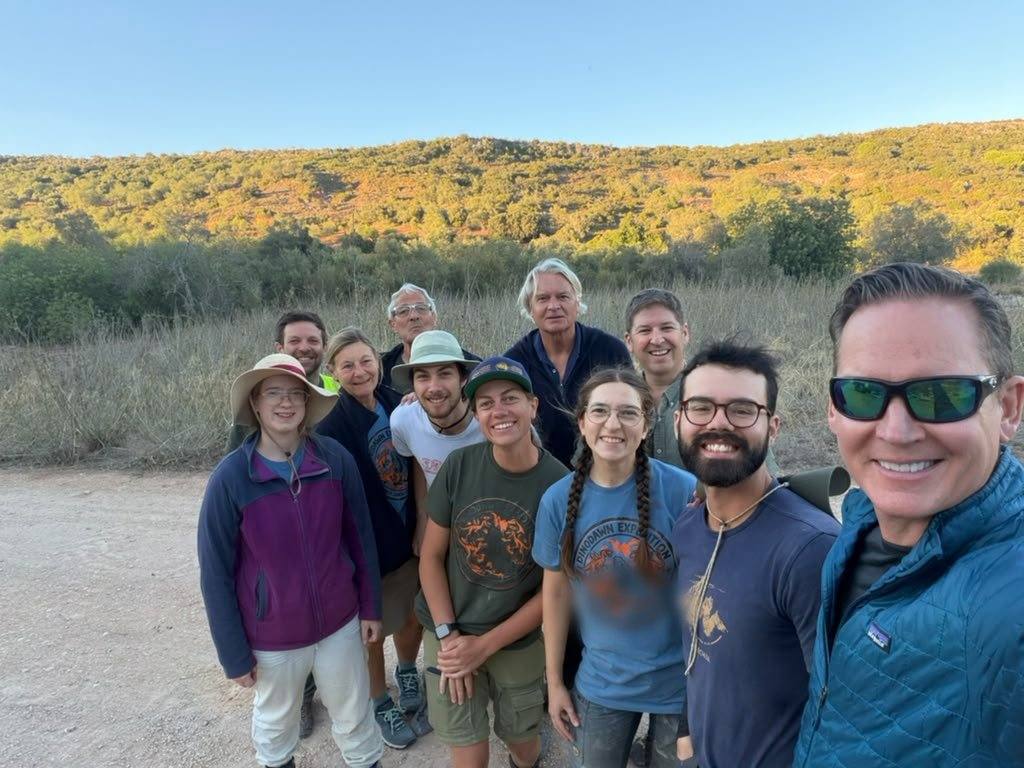 12 smiling team members gathered on a dirt track in front of a scrubby hillside in evening light