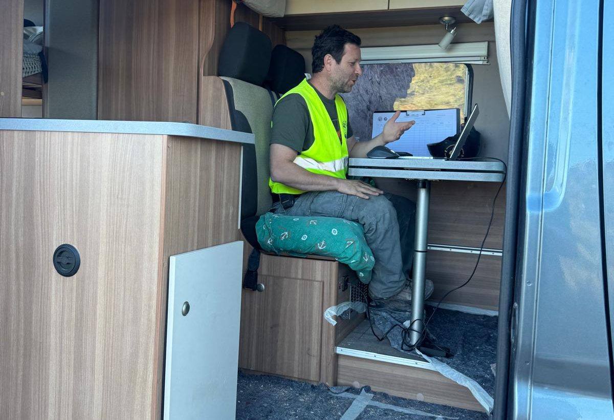 A man in a high visibility vest talking to a laptop at a small table inside a camper van