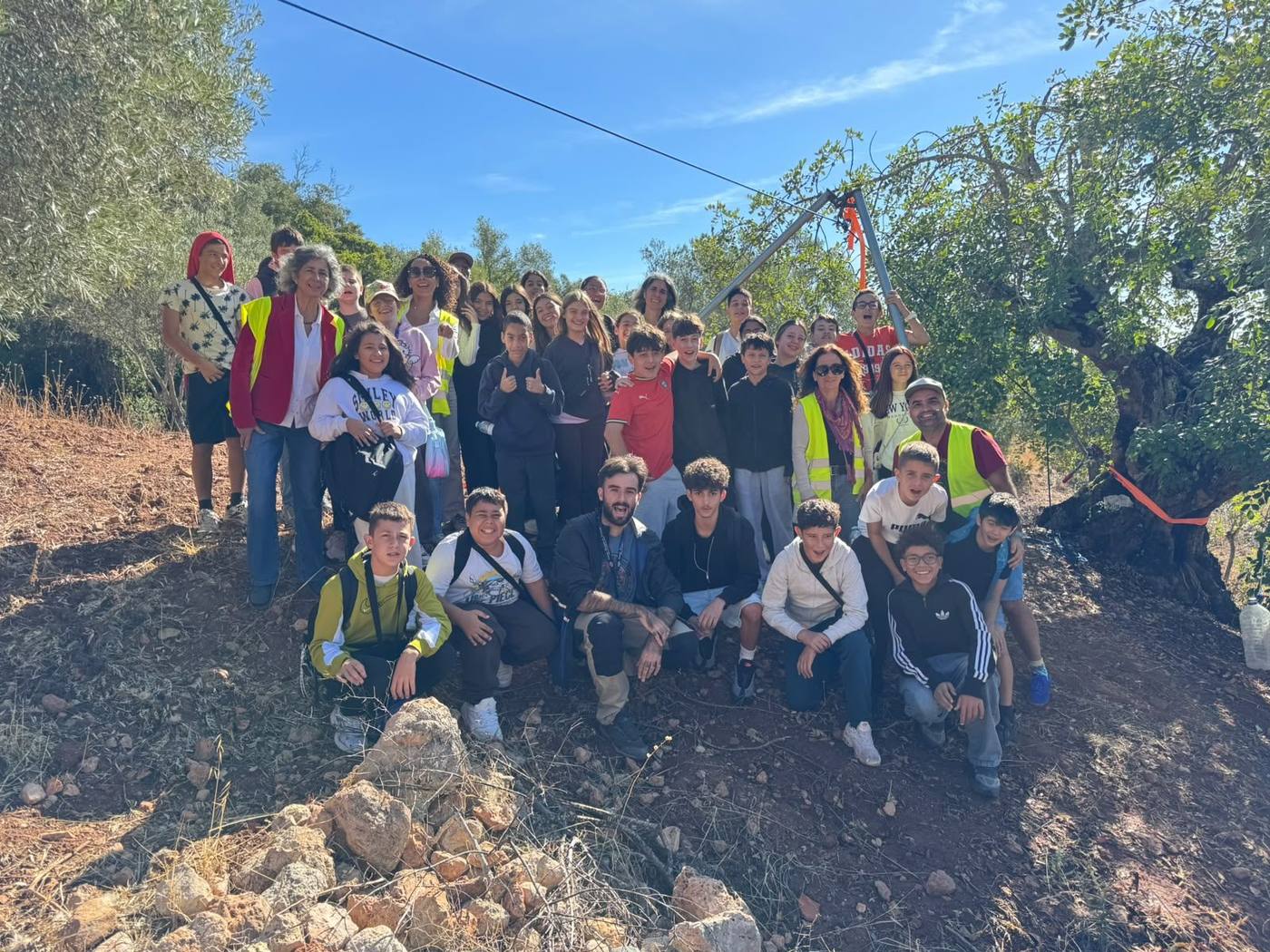 A large group of smiling students and teachers in high visibility vests posing on a hillside under olive trees