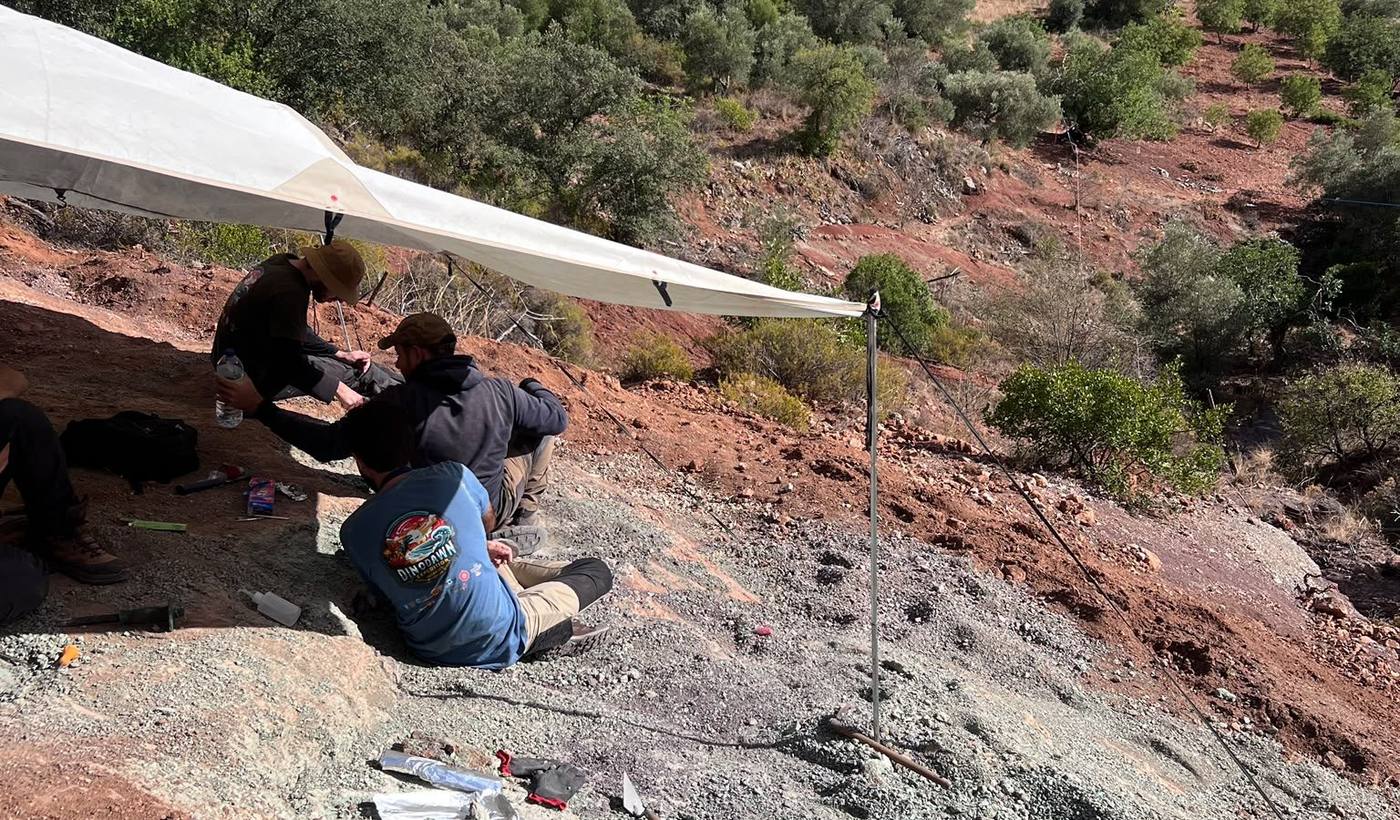 Team members sitting under a white tarp on a greenish rock slope, with wooded hills behind