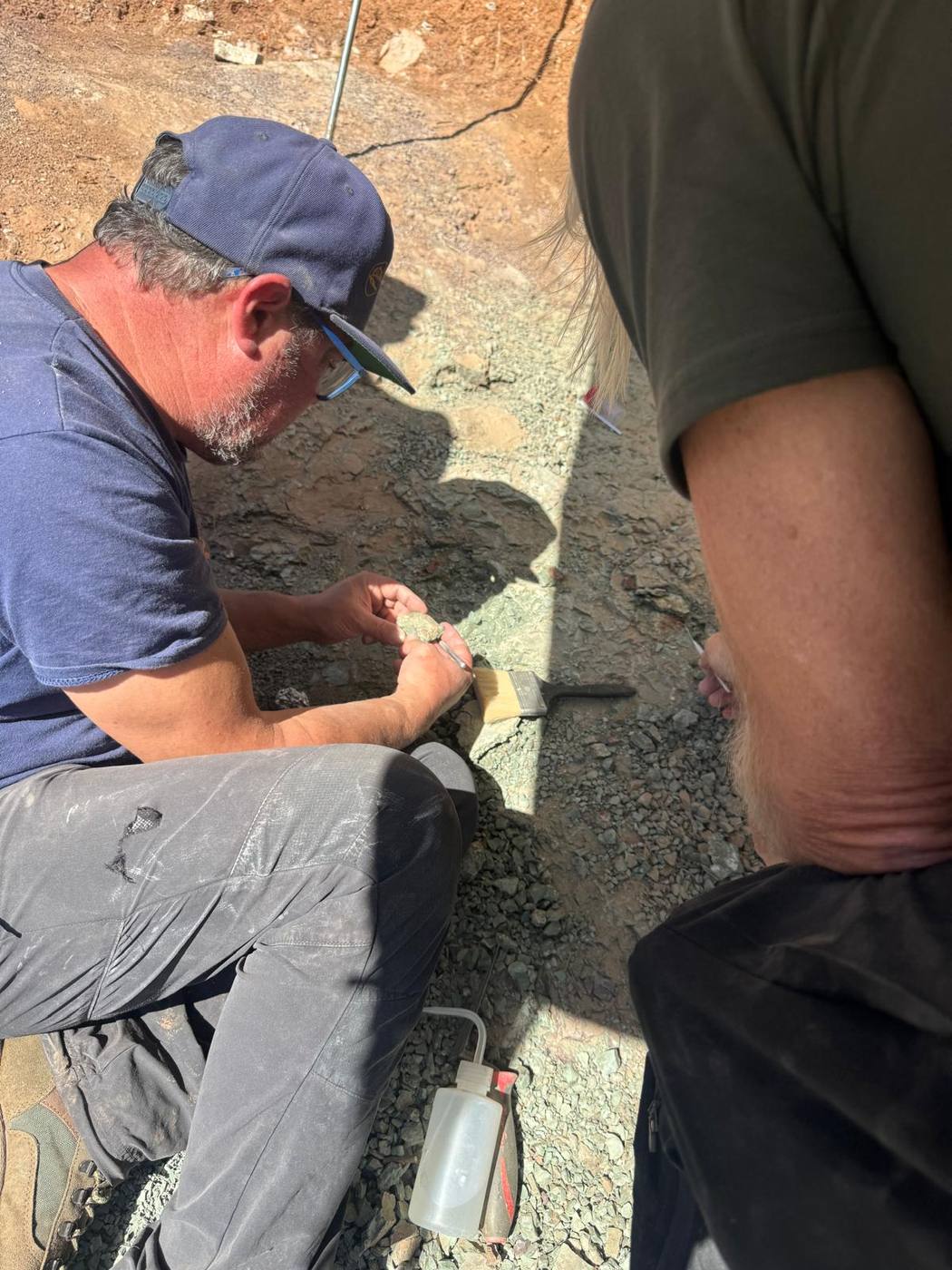 A team member in a cap crouching on the slope, brushing a small fossil piece held in his hand