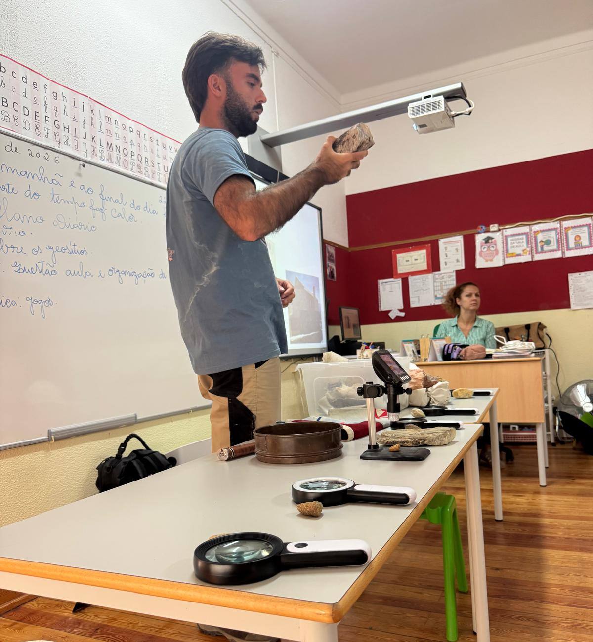 A man holding up a rock in a classroom, with magnifying glasses, a digital microscope and fossils on a table in front of him