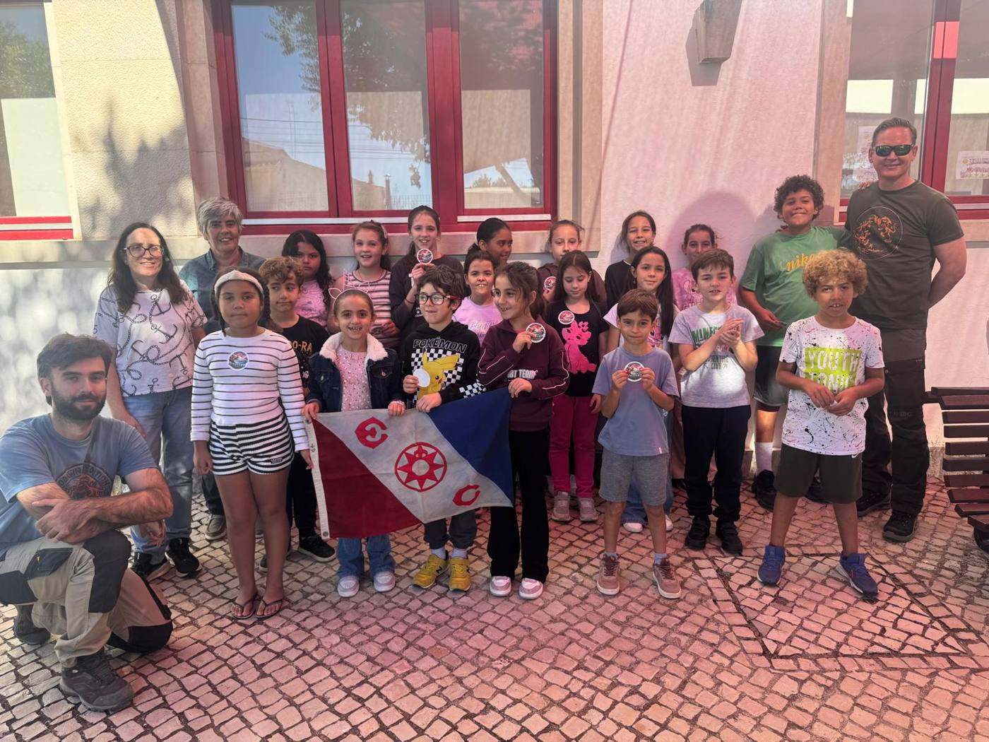A group of smiling students and adults outside a school with red window frames, 2 children holding a red, white and blue flag