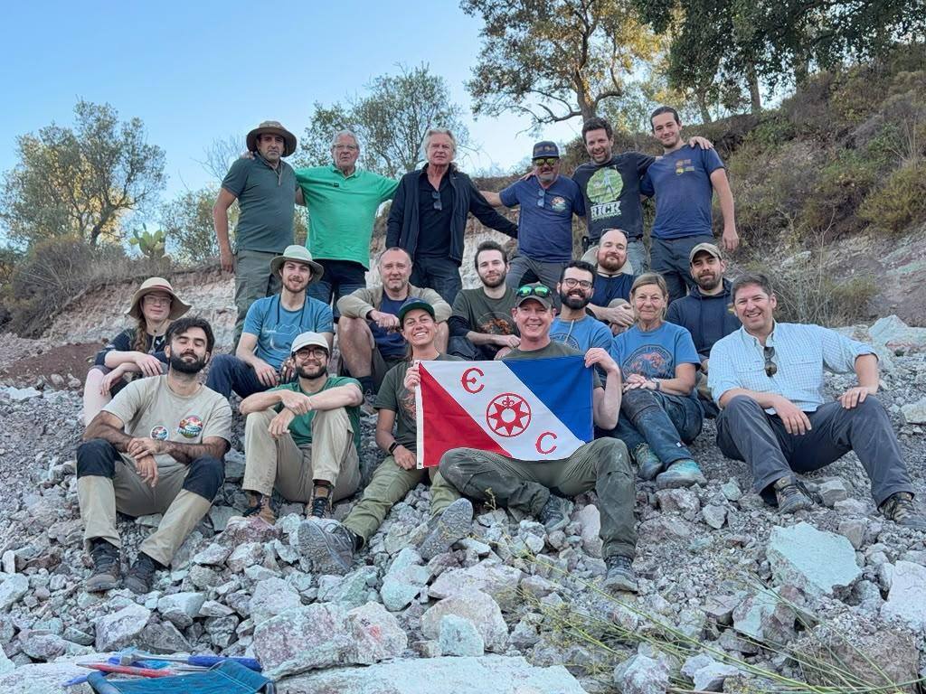 About 20 smiling team members sitting and standing on a rocky slope, 2 holding a red, white and blue flag