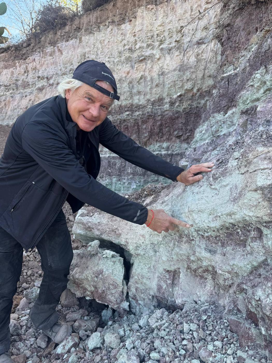 A smiling man in a dark cap pointing at a fossil in a large gray green block below a banded rock face