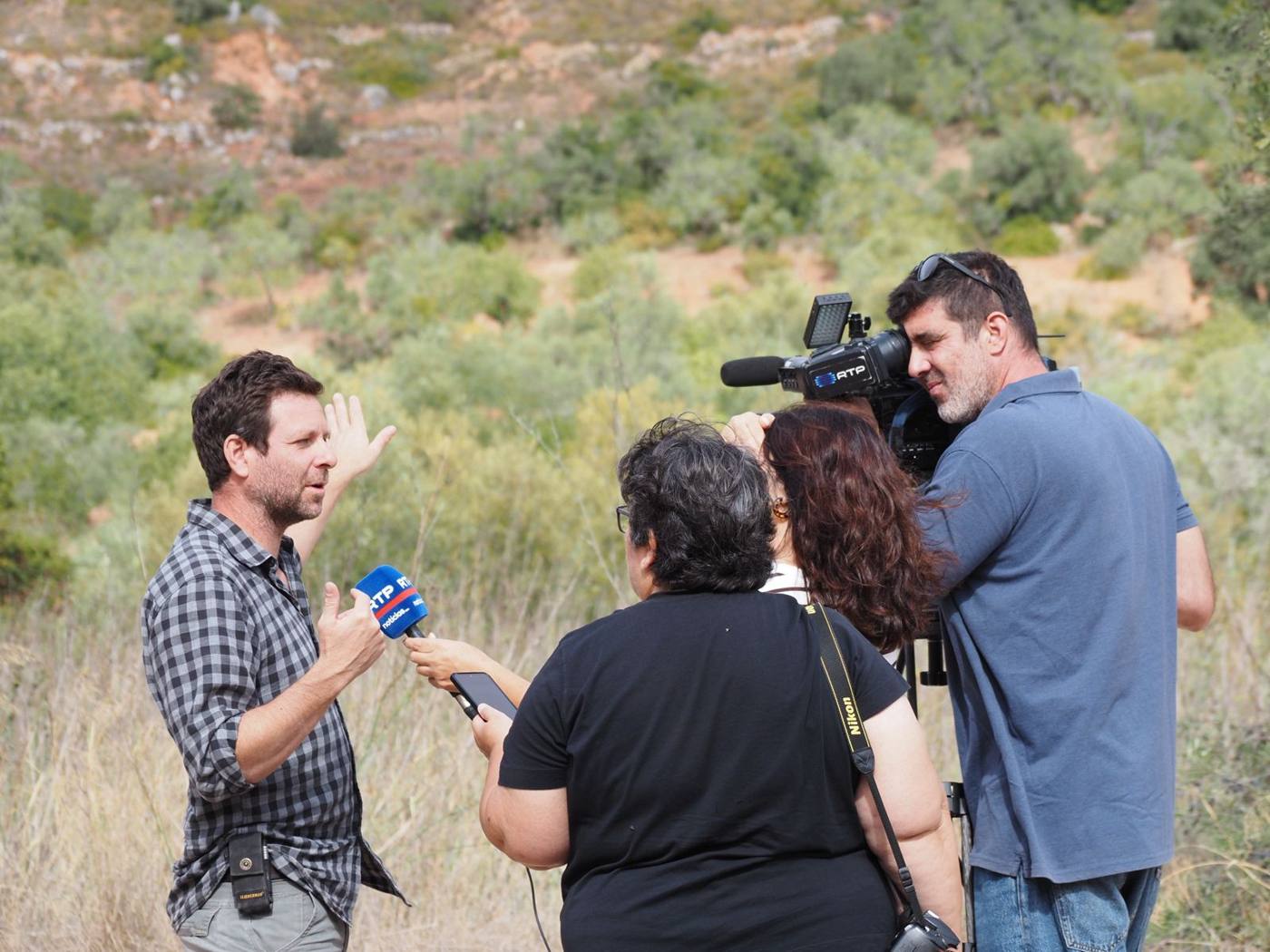 A man in a checked shirt gesturing as he talks into an RTP microphone, with a cameraman filming