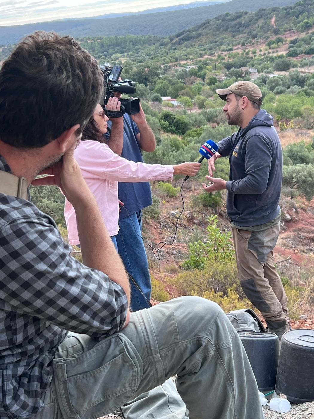 A man in a cap gesturing as he talks into an RTP microphone held by a journalist, with a cameraman filming and a team member watching