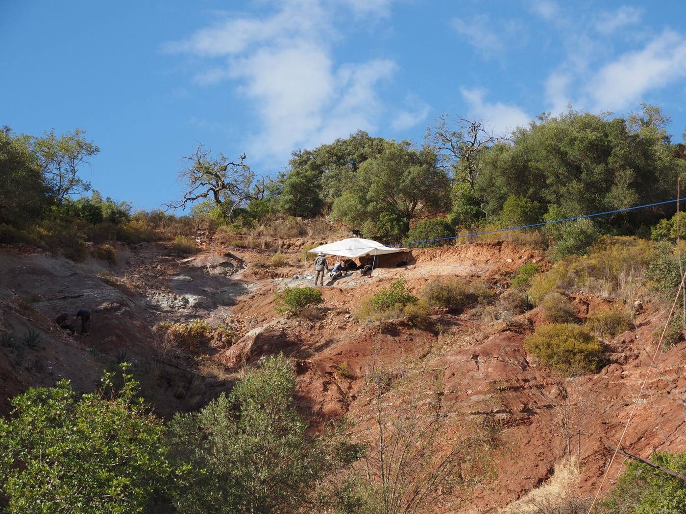A white tarp shading team members on a steep red and gray hillside below trees and a blue sky