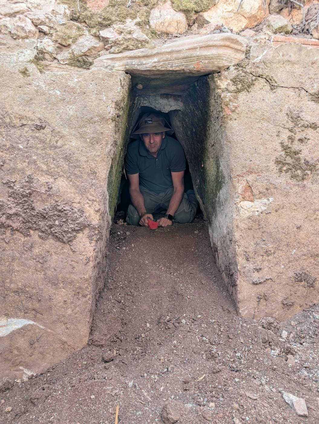 A man in a bush hat kneeling inside a narrow stone well entrance capped with a flat slab