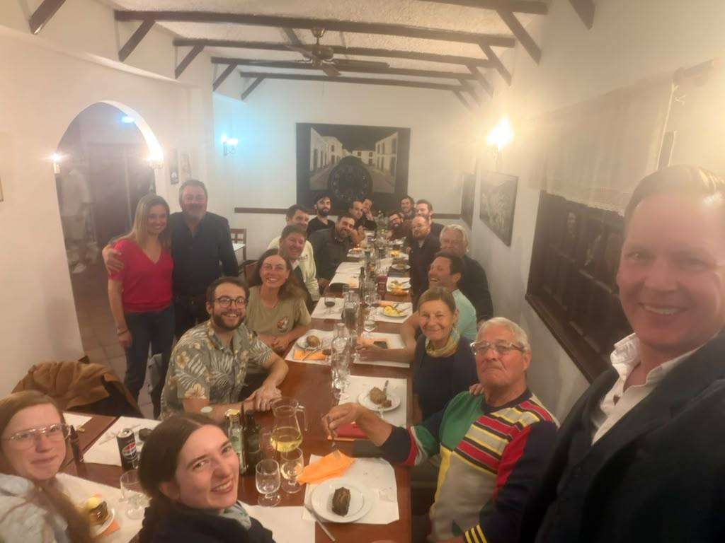 About 20 smiling team members around a long dinner table in a restaurant with white walls and wooden beams