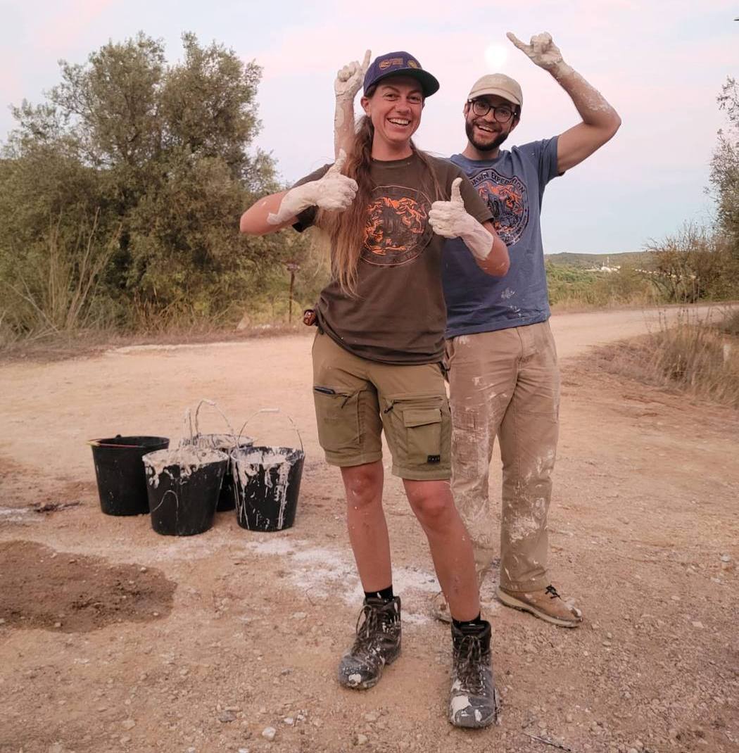 2 smiling team members with plaster covered hands and arms giving thumbs up beside plaster splashed buckets on a dirt track