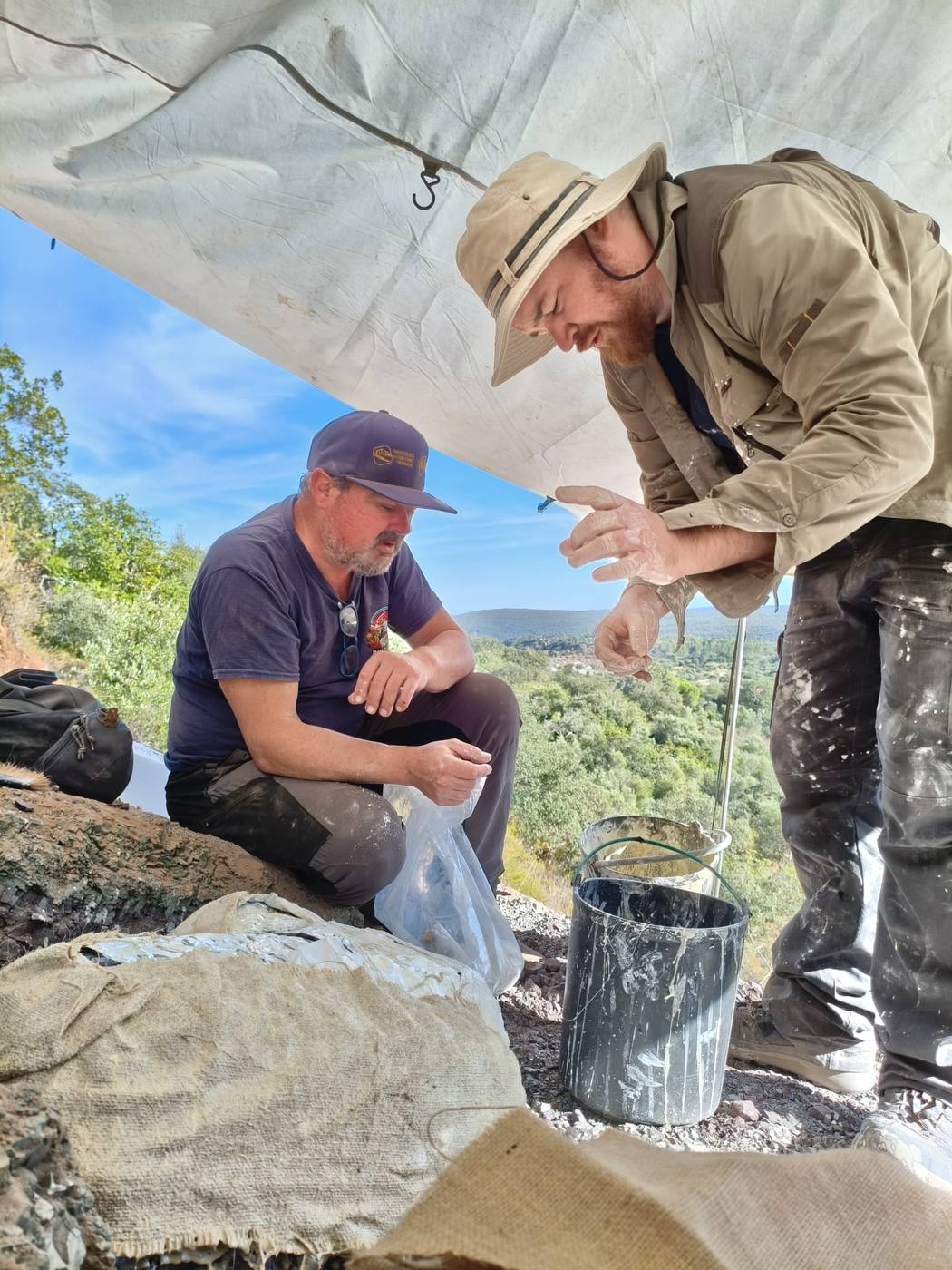 2 team members under a white tarp on a hillside, one crouched with a plastic bag, the other with plaster covered hands beside a bucket