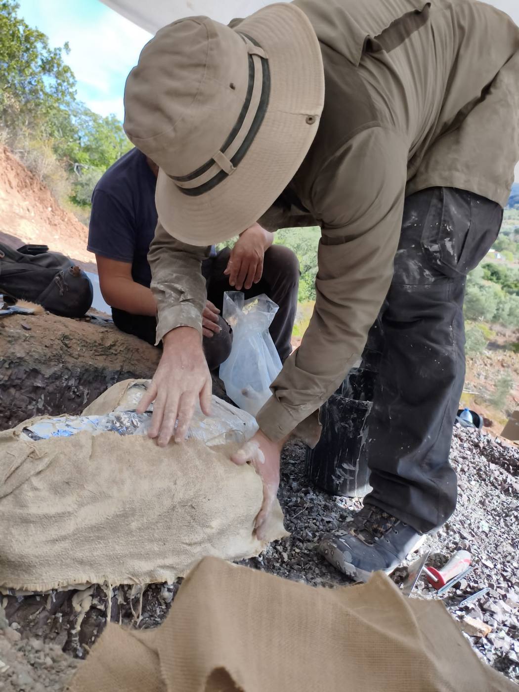A team member in a sun hat pressing burlap over a foil wrapped block on a gravel slope