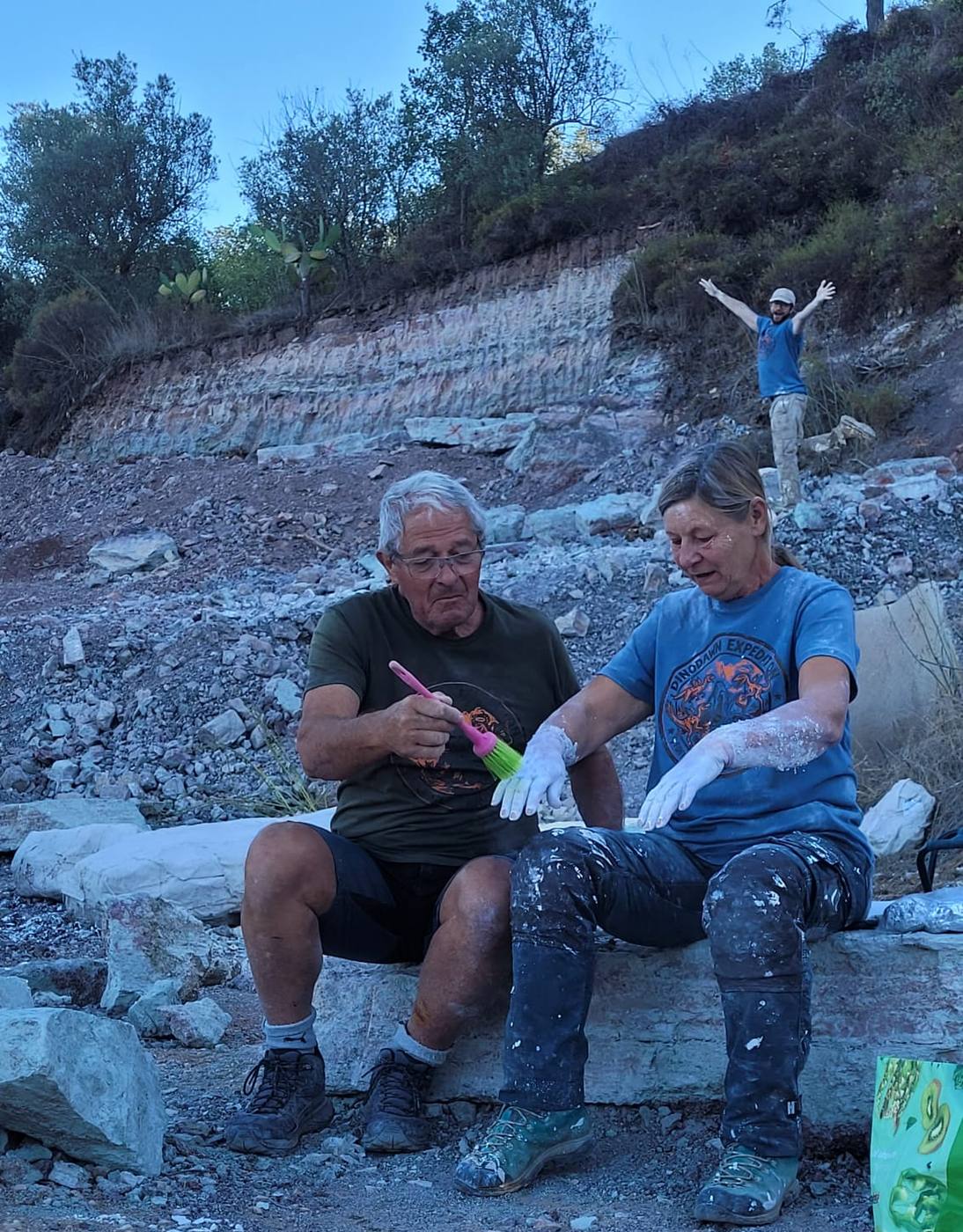 2 team members sitting on a block, one brushing plaster off the other’s hands with a pink brush, while a third throws his arms up on the slope behind