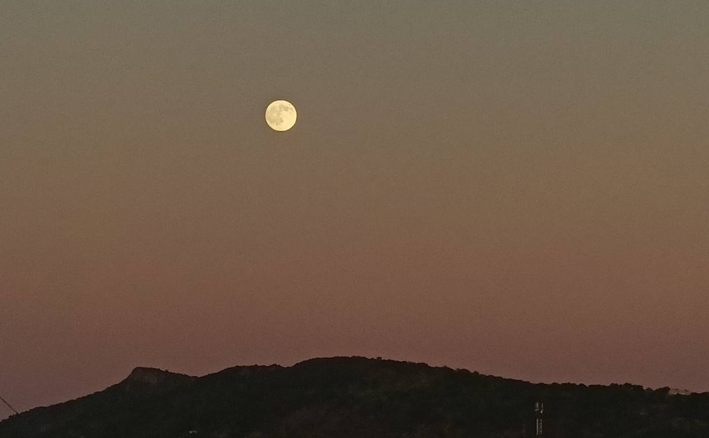 A pale full moon in a pink and violet dusk sky above a dark ridge line
