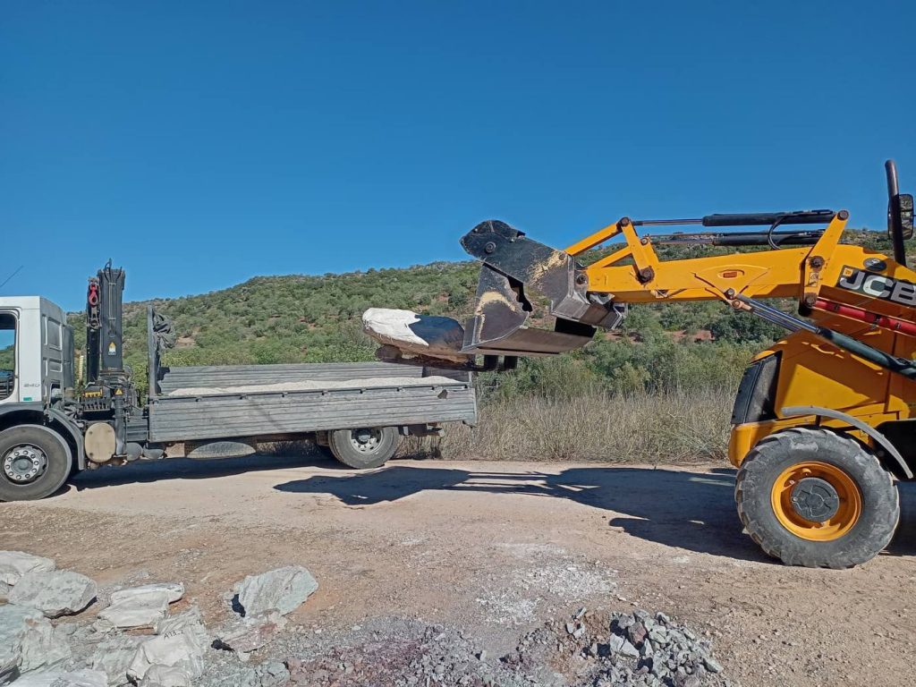 A yellow excavator lifting a plaster jacketed block in its bucket toward the bed of a flatbed truck