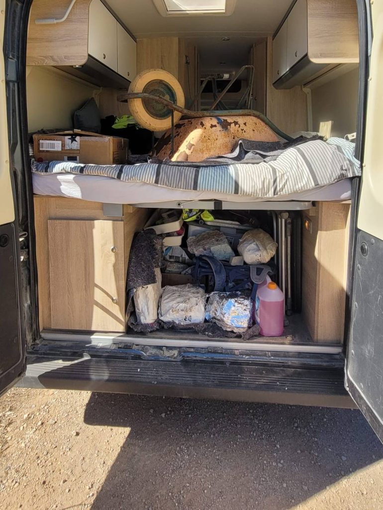 The open back of a camper van, with plaster jackets packed beneath the bed and a wheelbarrow on top