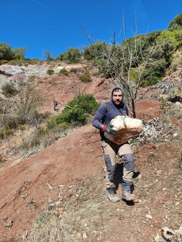 A team member in a hooded sweatshirt carrying a plaster jacket in his arms down a steep red hillside
