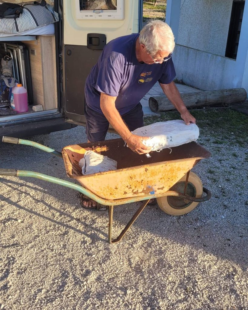 A team member lifting a labeled plaster jacket from a rusty wheelbarrow beside an open camper van