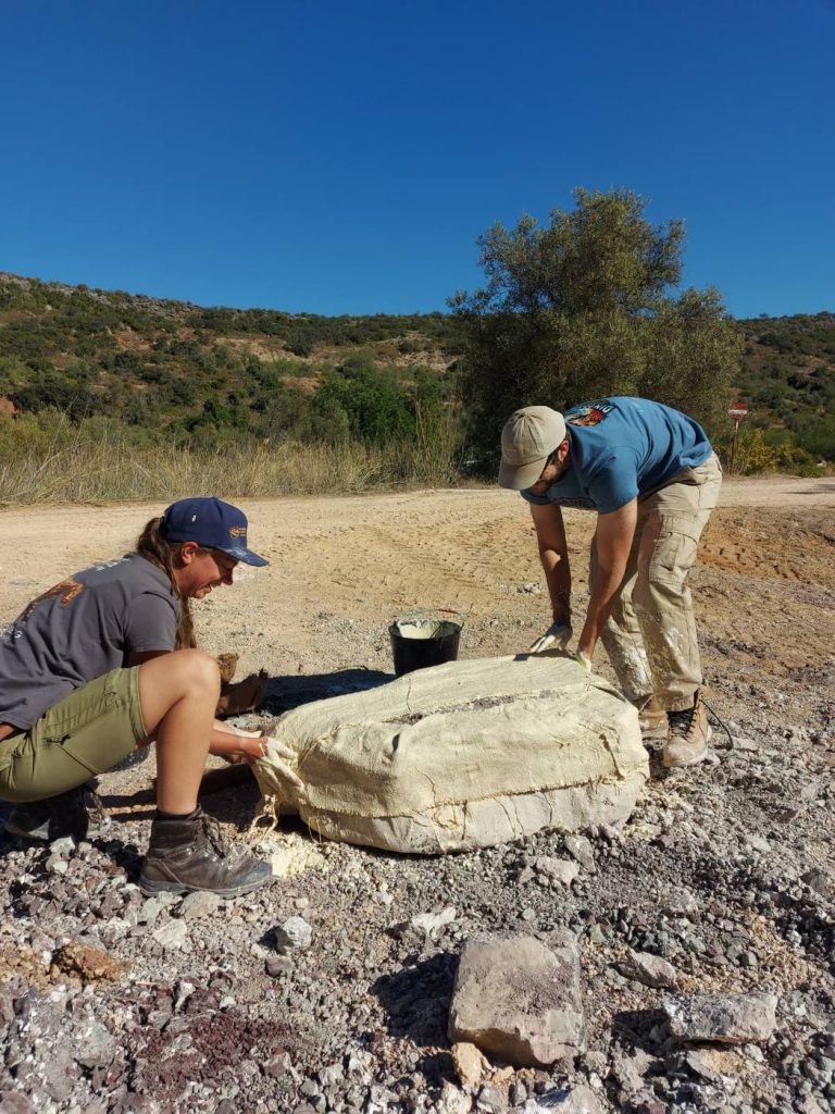 2 team members smoothing wet plaster bandages over a large block on a gravel track under a blue sky