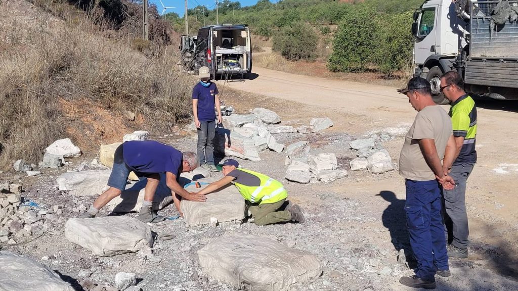 Team members kneeling over a plaster jacketed block on a gravel track, surrounded by more labeled jackets, while 2 men watch and a truck waits behind