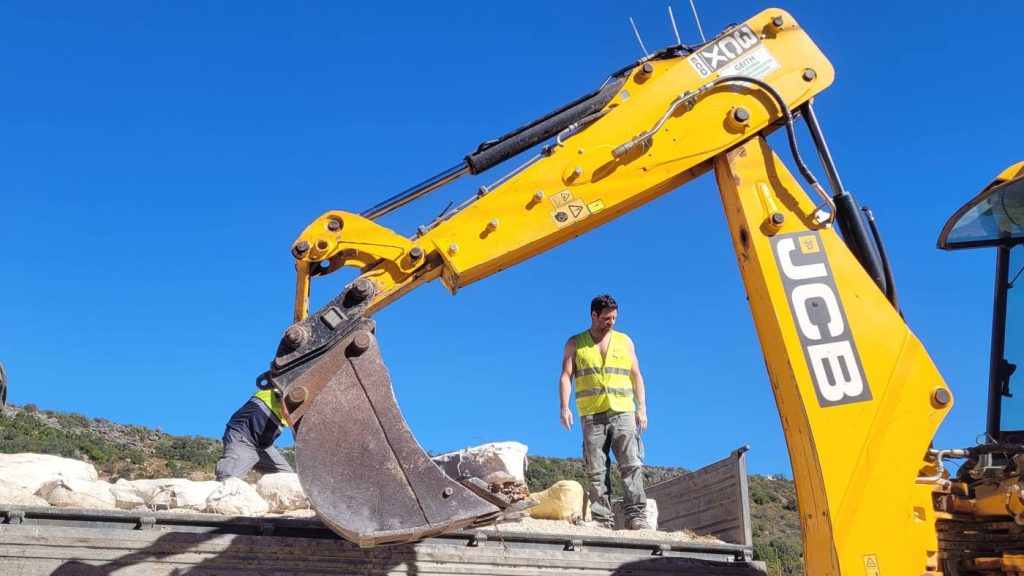 A team member in a high visibility vest standing on a truck bed among plaster jackets, beneath the arm of a yellow excavator