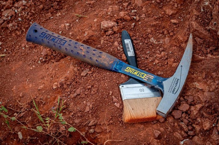 Rock hammer and brush lying on red Algarve soil at the dig
